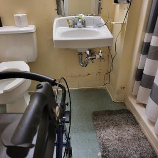 A view of an accessible bathroom with a wheelchair, featuring a toilet, sink, and shower area.