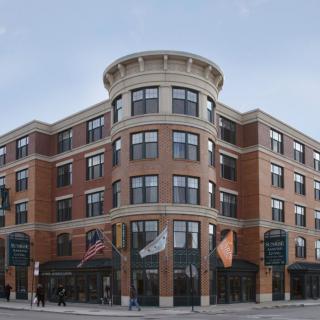 A modern, multi-story brick building situated at a street corner, featuring large windows and various flags displayed outside.
