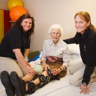 A caregiver assisting an elderly woman with therapy exercises, supported by a second caregiver.