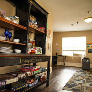 A cozy room with shelves filled with board games and kitchenware, illuminated by daylight through a large window.