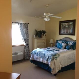 A cozy bedroom featuring a bed with blue and white bedding, a dresser, a window with curtains, and a ceiling fan.