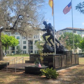A memorial statue surrounded by greenery, featuring the American flag in the background.