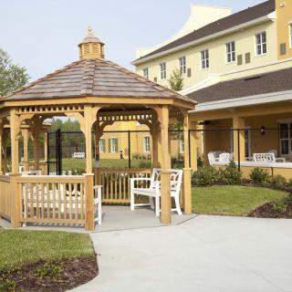 A wooden gazebo in front of a large yellow building surrounded by a landscaped garden.