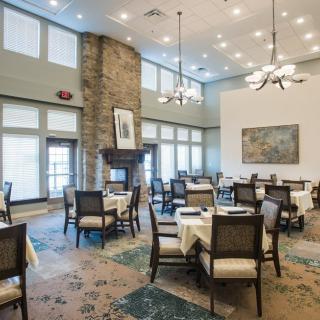 A spacious, elegantly decorated dining room featuring multiple tables and chairs, a stone fireplace, and large windows providing natural light.