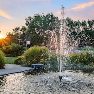 A tranquil park scene featuring a fountain with water jets, surrounded by lush greenery at sunset.