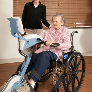 An elderly person in a wheelchair using an exercise machine with the assistance of a caregiver.