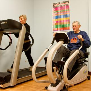 Two elderly individuals exercising on gym equipment, with one using a treadmill and the other on a stationary bike.