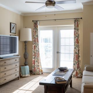 A cozy living room with a television, sofa, and coffee table, illuminated by natural light through large windows.