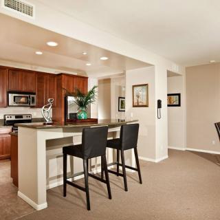 A modern apartment kitchen with an open layout, featuring wood cabinets and a breakfast bar with two bar stools.