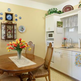 A cozy kitchen with a dining area, featuring a wooden table and chairs, a vase of flowers, and decorative wall elements.