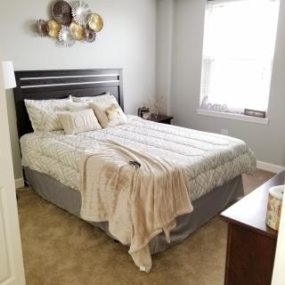 A cozy bedroom with a neatly made bed, decorative pillows, and a beige blanket draped over it. The room is brightened by natural light from a window.