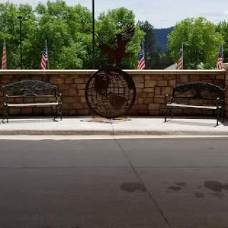 A peaceful outdoor setting featuring benches and a decorative sculpture against a backdrop of trees and flags.