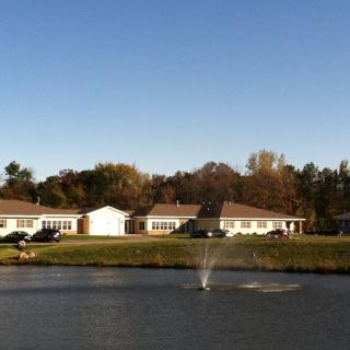 A serene lakeside view with a fountain, surrounded by houses and trees under a clear blue sky.