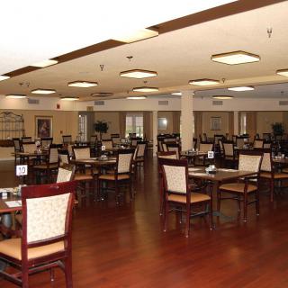 A spacious indoor restaurant dining area with empty tables and chairs, featuring wooden floors and overhead lighting.