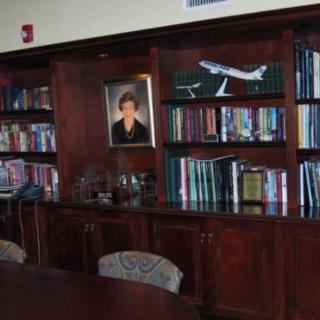 A wooden bookshelf filled with books, a model airplane, and a portrait hanging above.