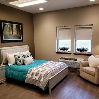 A cozy bedroom featuring a neatly made bed, an upholstered chair, and two windows adorned with blinds. The room is decorated with a nature-themed artwork.