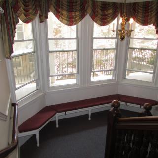 A cozy indoor space with a bay window, featuring built-in red-cushioned seating and decorative curtains overlooking the staircase.