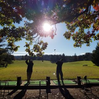 Two people practicing at a driving range under the sunlight.