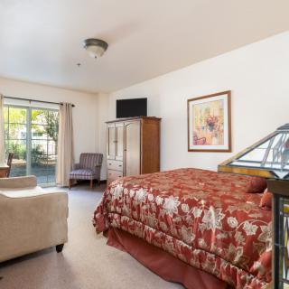 A cozy bedroom featuring a bed with a floral cover, a window with curtains, and various furniture pieces including a chair and dresser.
