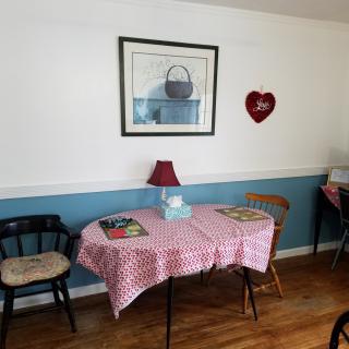 A cozy dining area with a table covered by a patterned tablecloth, surrounded by chairs and decorated with wall art and a small lamp.