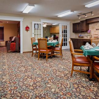 A cozy dining area with a carpeted floor, set with tables and chairs, leading to a kitchen space.