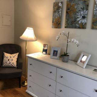 A cozy corner featuring a white dresser, upholstered chair, standing lamp, and decorative elements.