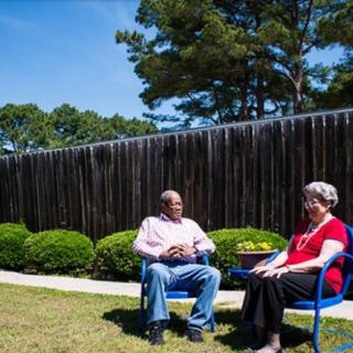 Two people sitting on chairs in a garden, having a conversation under a clear blue sky.