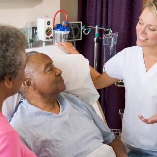 A nurse talking to a patient in a hospital room, with a visitor standing nearby.