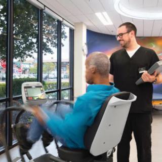 A healthcare professional assists a person exercising on a machine in a gym setting.
