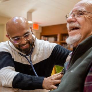 A healthcare professional using a stethoscope to check an elderly patient's heart, both smiling during the interaction.