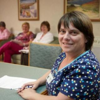 A smiling woman seated at a table with papers in front of her, surrounded by others in an office setting.
