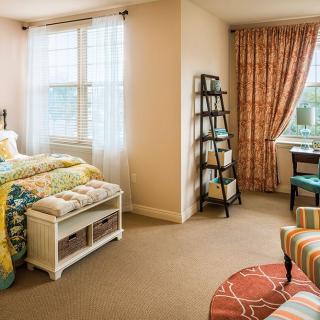 A cozy bedroom with colorful bedding, a small bookshelf, and natural light from large windows.