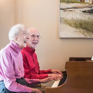 An elderly couple enjoys playing the piano together indoors, sharing a joyful moment.