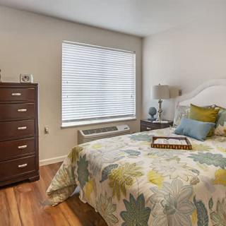 A cozy bedroom with a floral bedspread, wooden dresser, and window with blinds.