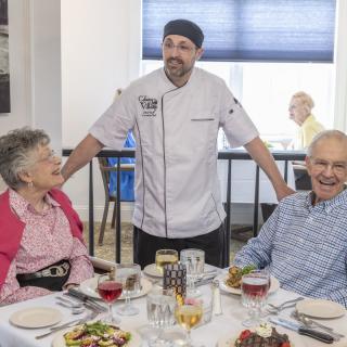 A chef standing with two senior patrons enjoying a meal at a restaurant.