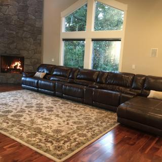 A cozy living room featuring a large leather sectional sofa, a warm fireplace, and expansive windows overlooking greenery.