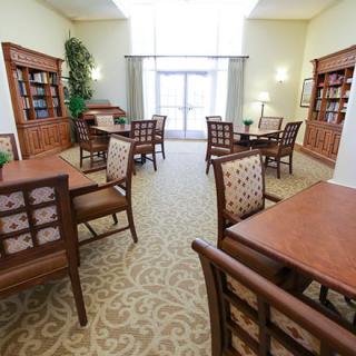 A well-lit library interior with wooden tables and chairs, surrounded by bookshelves.