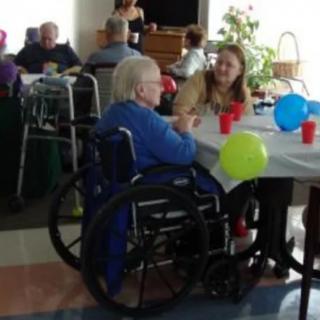 A social gathering at a nursing home with residents and staff interacting around a table decorated with colorful balloons.