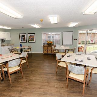 A well-lit dining area with multiple tables and chairs arranged neatly, featuring a kitchenette and decorative elements on the walls.