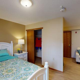 A cozy bedroom featuring a bed with colorful bedding, a bedside table with a lamp, and an open wardrobe.