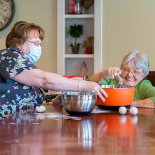 Two women engaged in a cooking activity together, both wearing masks in a cozy kitchen setting.