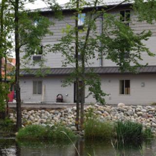 A serene view of a two-story building by a calm body of water, surrounded by lush greenery.