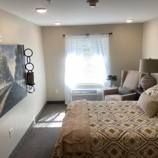 A cozy bedroom with a neatly made bed, armchair, and bright natural light coming through the window.