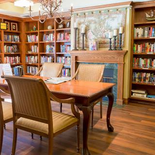 A cozy library room featuring a wooden table and chairs, surrounded by bookshelves and a fireplace.