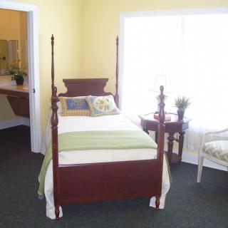 A cozy bedroom featuring a four-poster bed, a side table, and an upholstered chair, with natural light streaming in through the window.