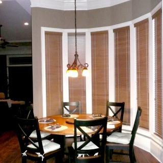 A cozy dining area with a round table, four chairs, and a pendant light fixture, surrounded by tall windows with blinds.