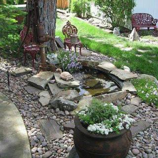 A serene backyard garden with a small pond surrounded by stones, lush greenery, and comfortable chairs for relaxation.