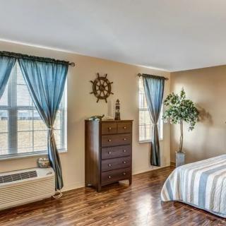 A cozy bedroom with wooden flooring, featuring a dresser, bed, and large windows adorned with blue curtains.