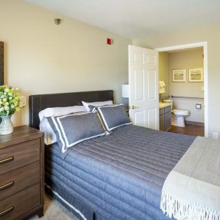 A cozy bedroom with a neatly made bed, nightstand with flowers, and an open door leading to the bathroom.