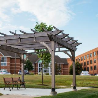 An outdoor area with a wooden pergola and benches, surrounded by grassy lawns and buildings.